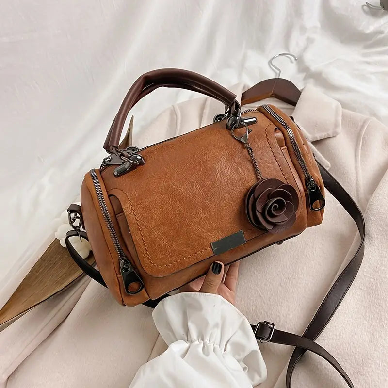 Brown leather handbag with floral detail held by a person wearing a white sleeve.