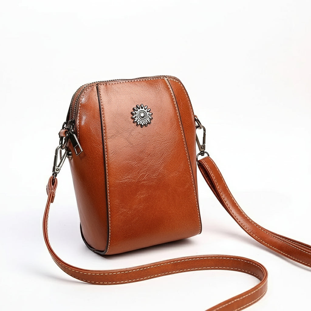 Brown leather crossbody bag with decorative emblem on a beige and orange background
