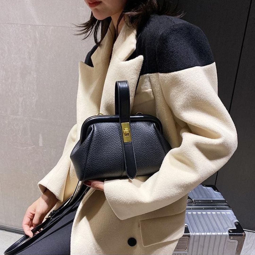 Person holding a black handbag wearing a beige and navy coat with a neutral background.